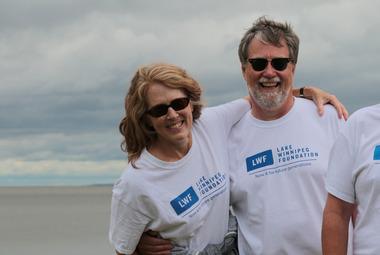 A woman standing with her arm around a man's shoulders, both standing in front of the lake, wearing LWF T-shirts and smiling.